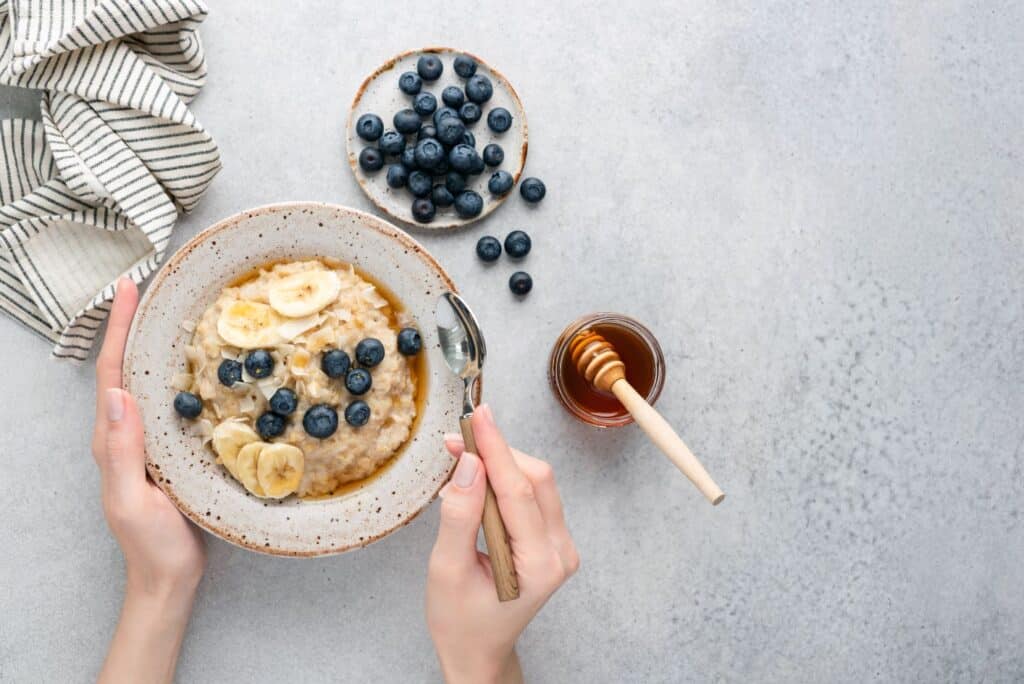 A bowl of oatmeal topped with sliced bananas and blueberries is held by two hands, one holding a spoon. A small bowl of blueberries and a jar of honey with a dipper are placed to the side. A striped cloth is on the upper left corner of a gray surface. MyFitnessPal Blog